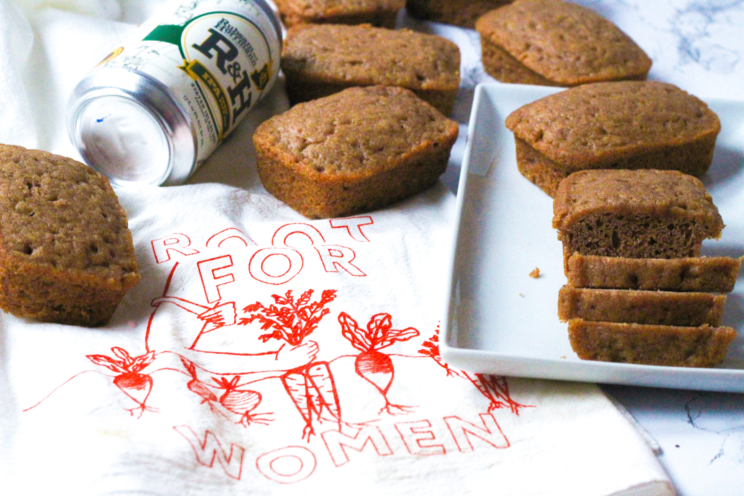 On the left side of the photo is a white dish towel that says Root for Women in red with root vegetables on it. In the upper left of the image on top of the towel above the words are some mini loaves of spiced beer bread and a can of R&H IPA Beer. On the right side of the photo is a white rectangular plate that slightly overlaps the towel. On the plate in the back is a mini loaf, and in the front is a loaf, the front half of which is sliced into three slices.