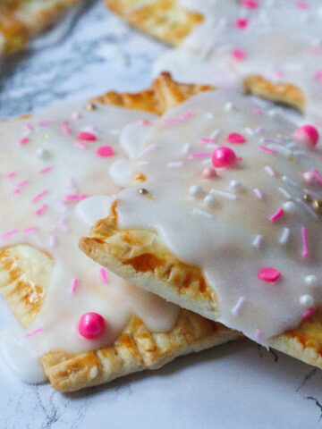 Close up of two strawberries & champagne poptarts, one sitting partially on top of the other on a white marbled surface. They are covered in white icing, and topped with pink sprinkles. You can see the edges of a couple more poptarts in the background.