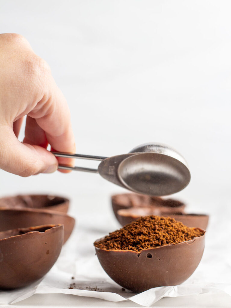 A hand pouring a spoon of espresso powder into a chocolate half-sphere shell.