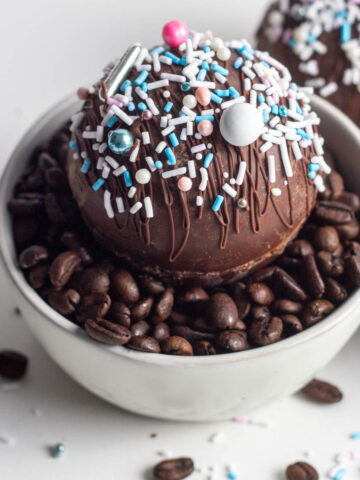 A hot mocha bomb covered in sprinkles sitting in a white bowl of coffee beans.