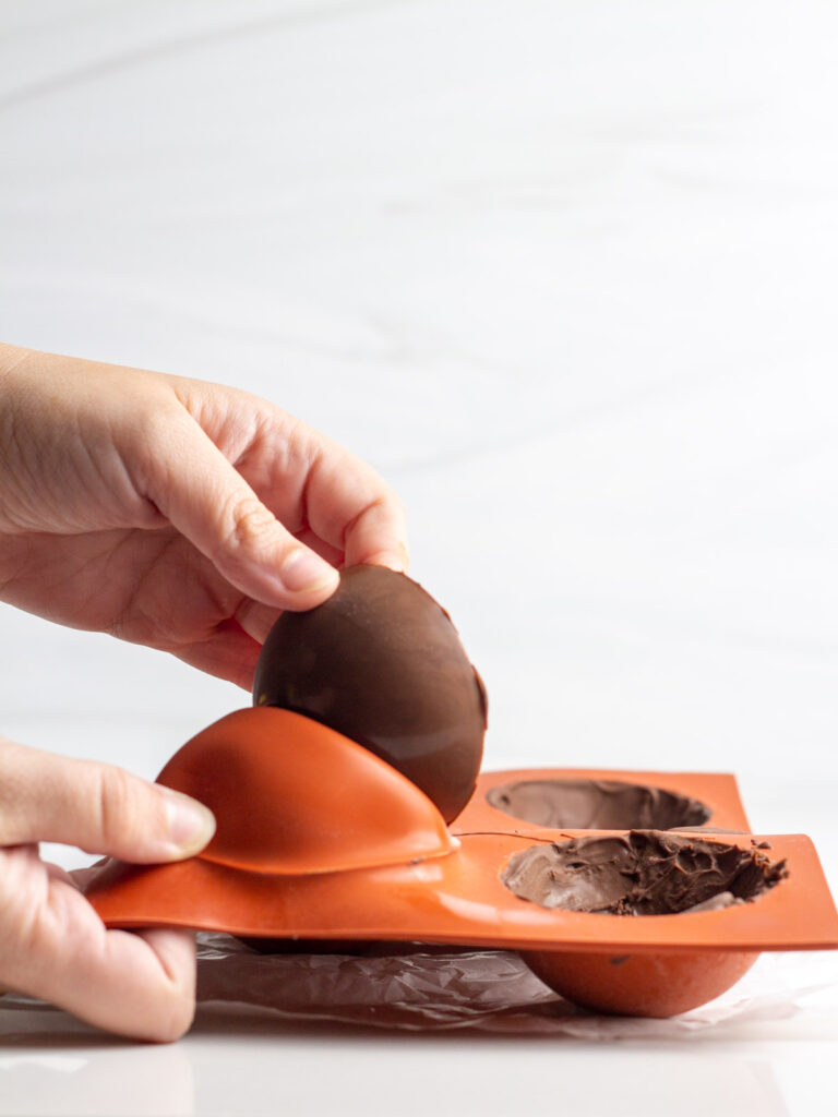 A hand peeling a hot chocolate bomb shell out of a red silicone mold.