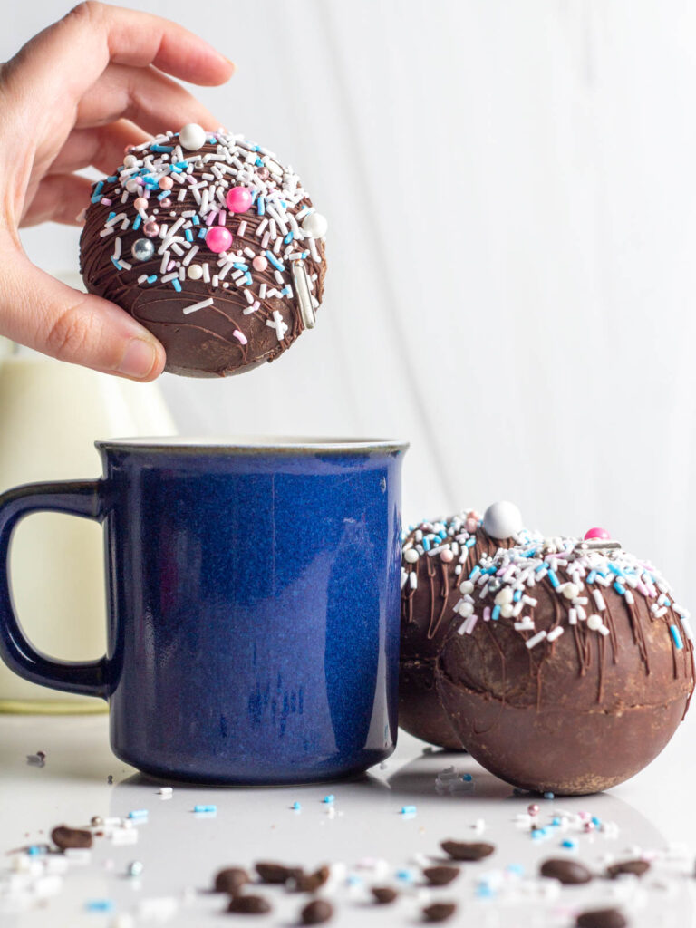 A hand holding a mocha hot chocolate bomb over a blue mug with more bombs next to the mug.