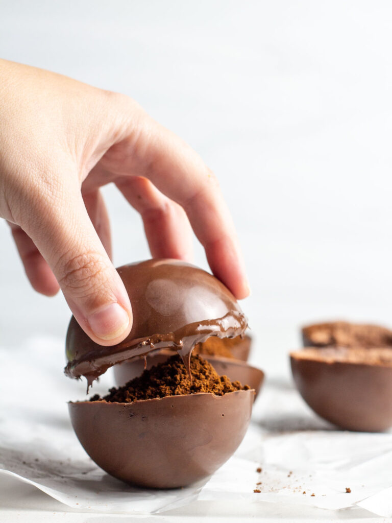 A hand placing an empty chocolate half-sphere on top of a filled half-sphere.