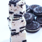 Side view of a stack of 3 pieces of cookies and cream fudge with a pile of oreos in the background, all on a white surface