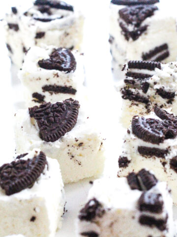 An angled view of a bunch of pieces of cookies and cream fudge (white cubes with pieces of Oreo throughout) on a white surface.