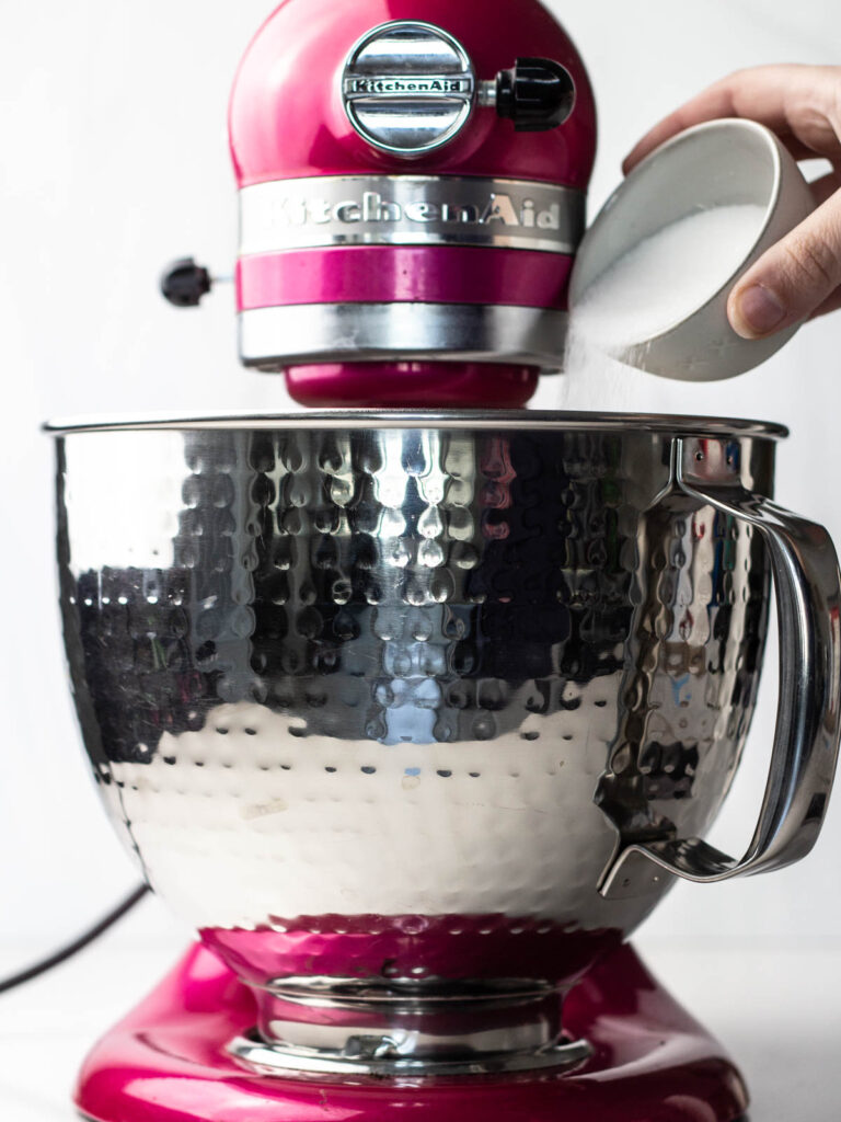 A hand pouring a small bowl of sugar into a textured steel mixing bowl on a pink kitchen aid mixer.