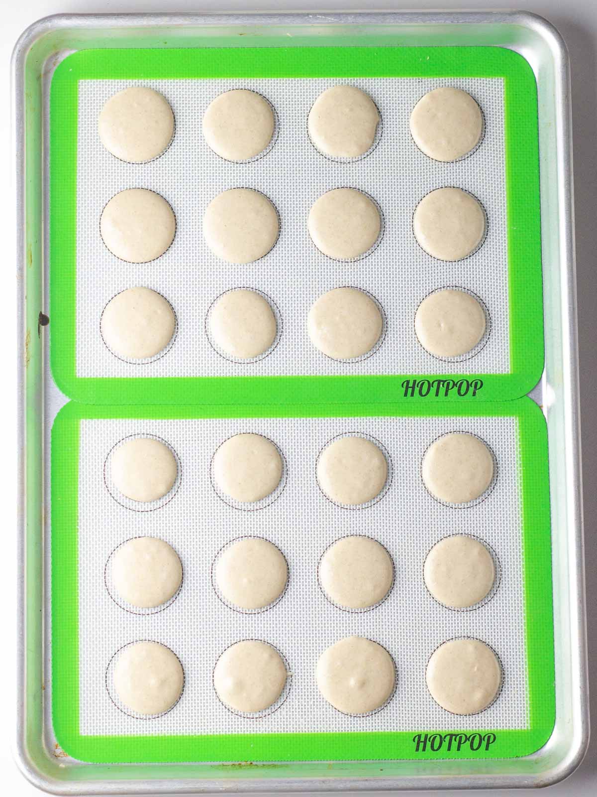 Two lined quarter baking sheets with macaron shells piped onto the silicone liners.
