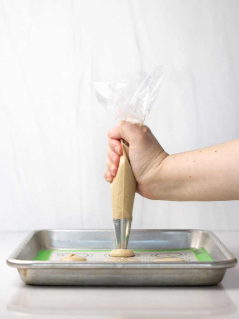 A hand holding a piping bag piping macaron batter onto a lined quarter sheet pan.