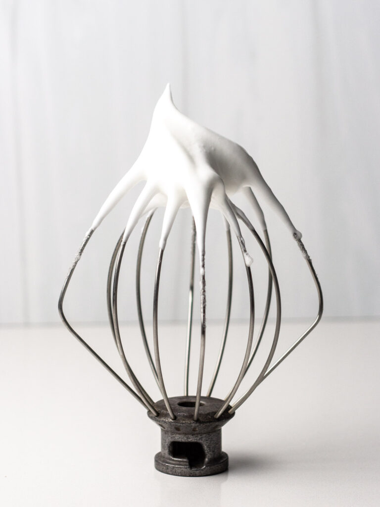 The whisk attachment from a stand mixer upside down with a peak of meringue sticking up from the whisk.