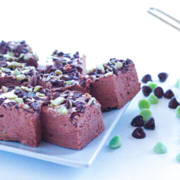 Side view of a white square plate of mint chocolate chip fudge on the left side of the frame. On the right side of the frame is a gold measuring cup overflowing with mint chocolate chips.