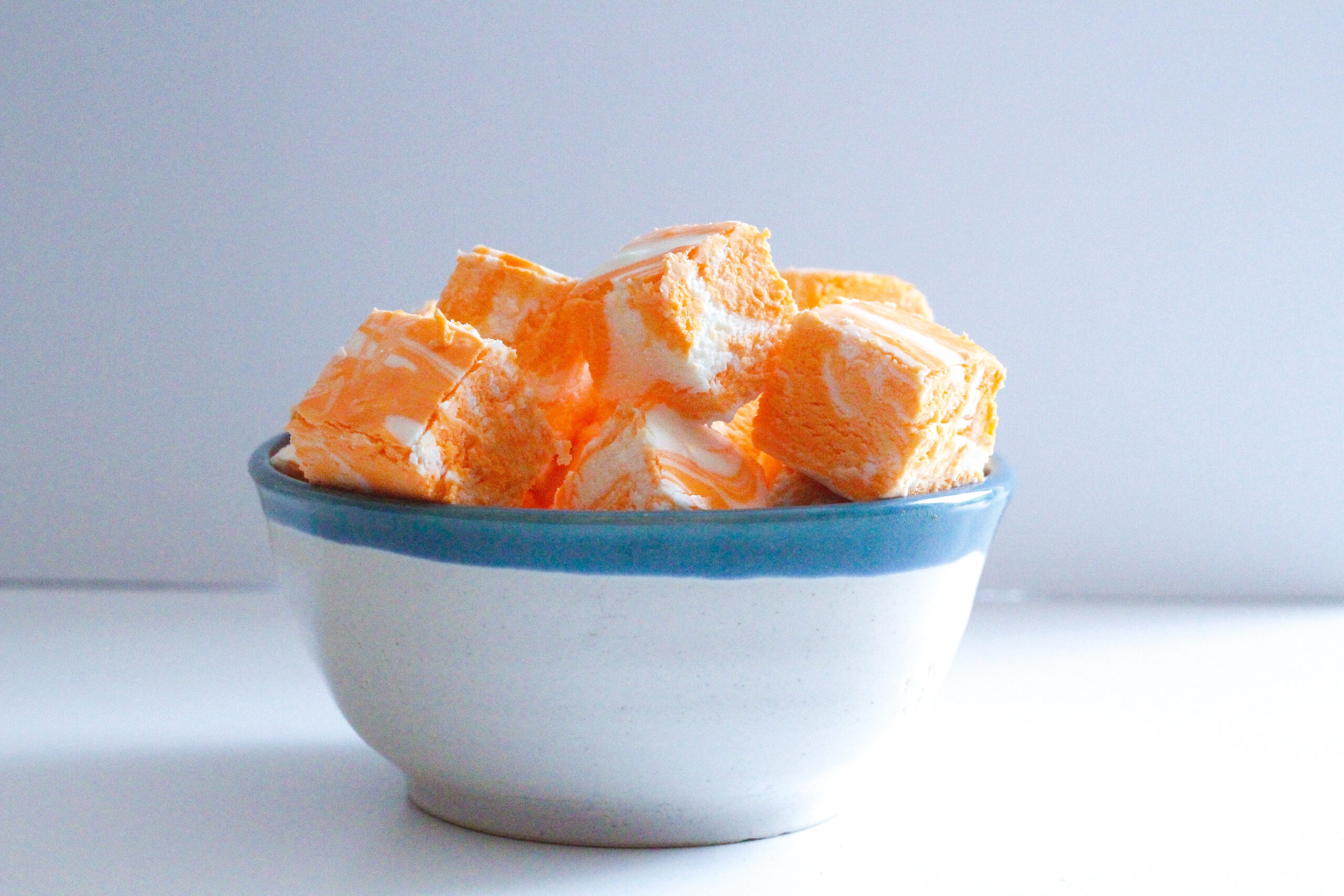 Side view of a white bowl with a blue rim filled with creamsicle fudge, orange and white swirled fudge cubes