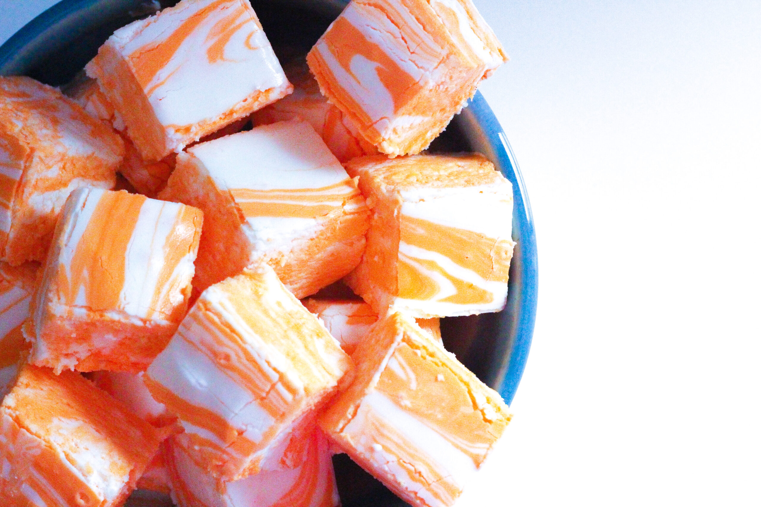 Top down view of creamsicle fudge - squares of swirled orange and white, sitting in a blue-rimmed bowl on a white surface.