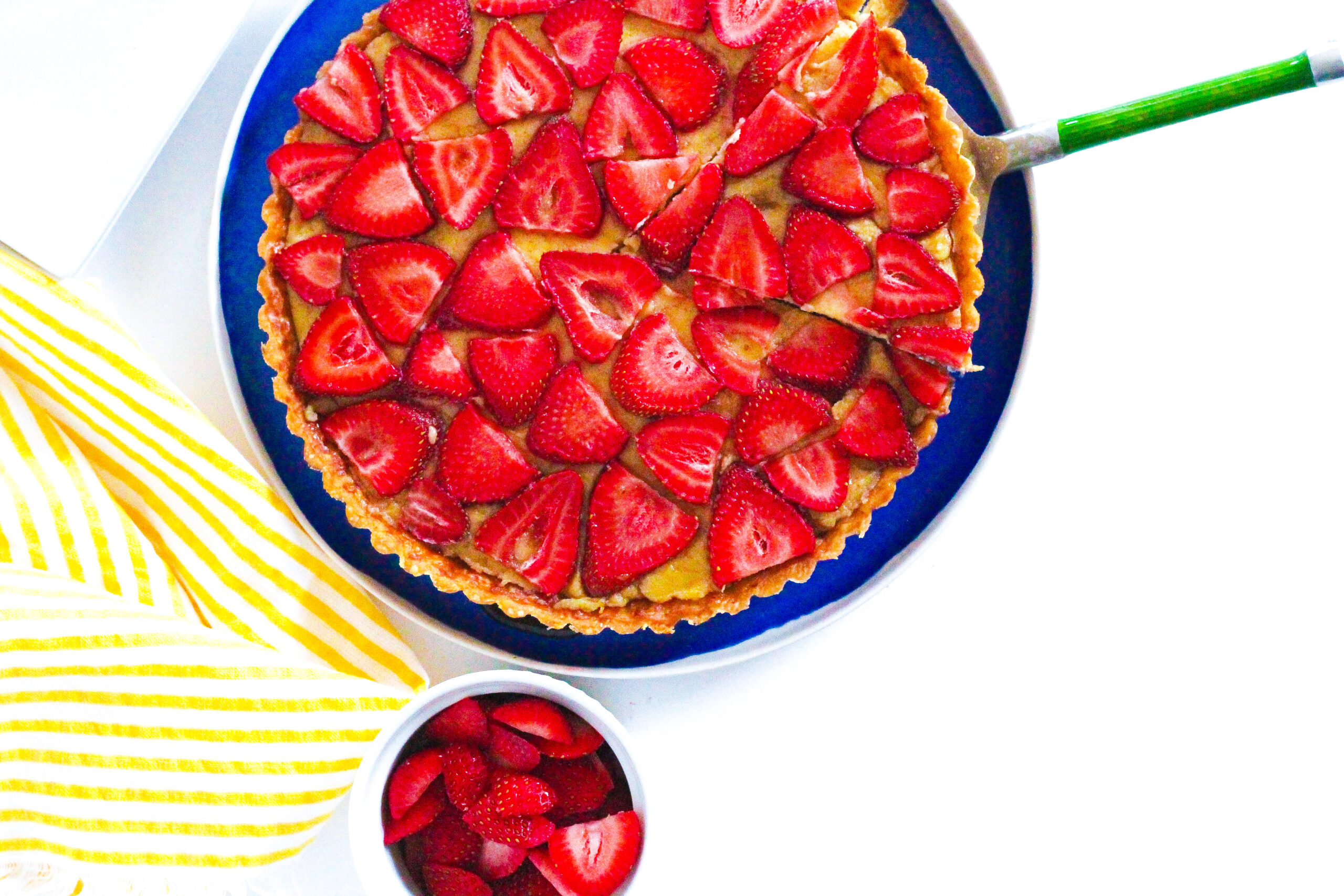 Top down view of a strawberry balsamic tart with the tart cut off of the top of the frame. A slice is cut out of the tart with a pie server under it, the green handle of the pie server sticking out to the right of the frame. below the tart is a small bowl of sliced strawberries, and to the left is a yellow striped towel. The tart is on a blue plate, and the scene is on a white surface.