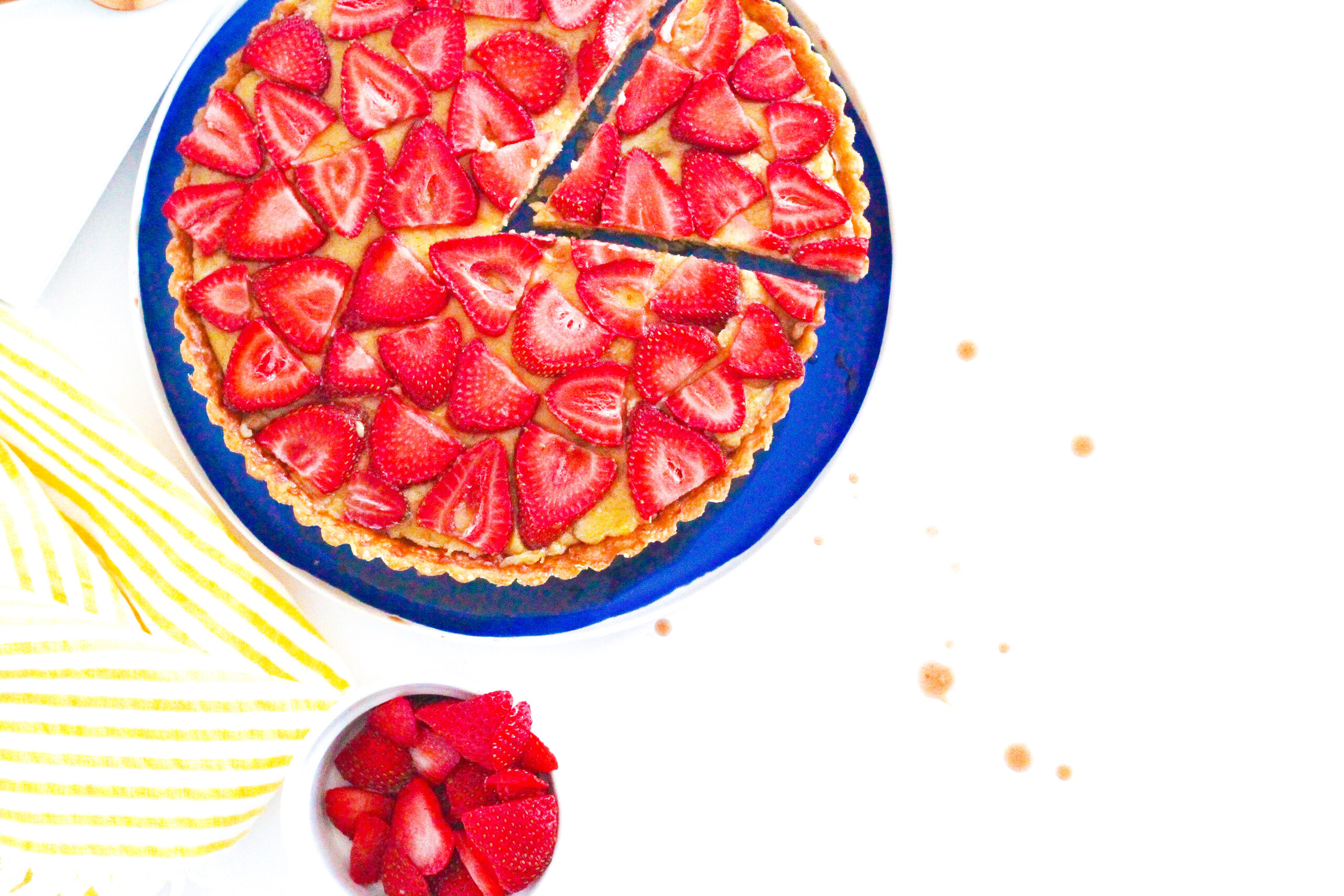 A slice slightly shifted out of a balsamic strawberry and basil tart with a bowl of strawberries below the tart and a yellow striped napkin to the left of the tart. To the right of the frame are splatters of balsamic on a white surface.