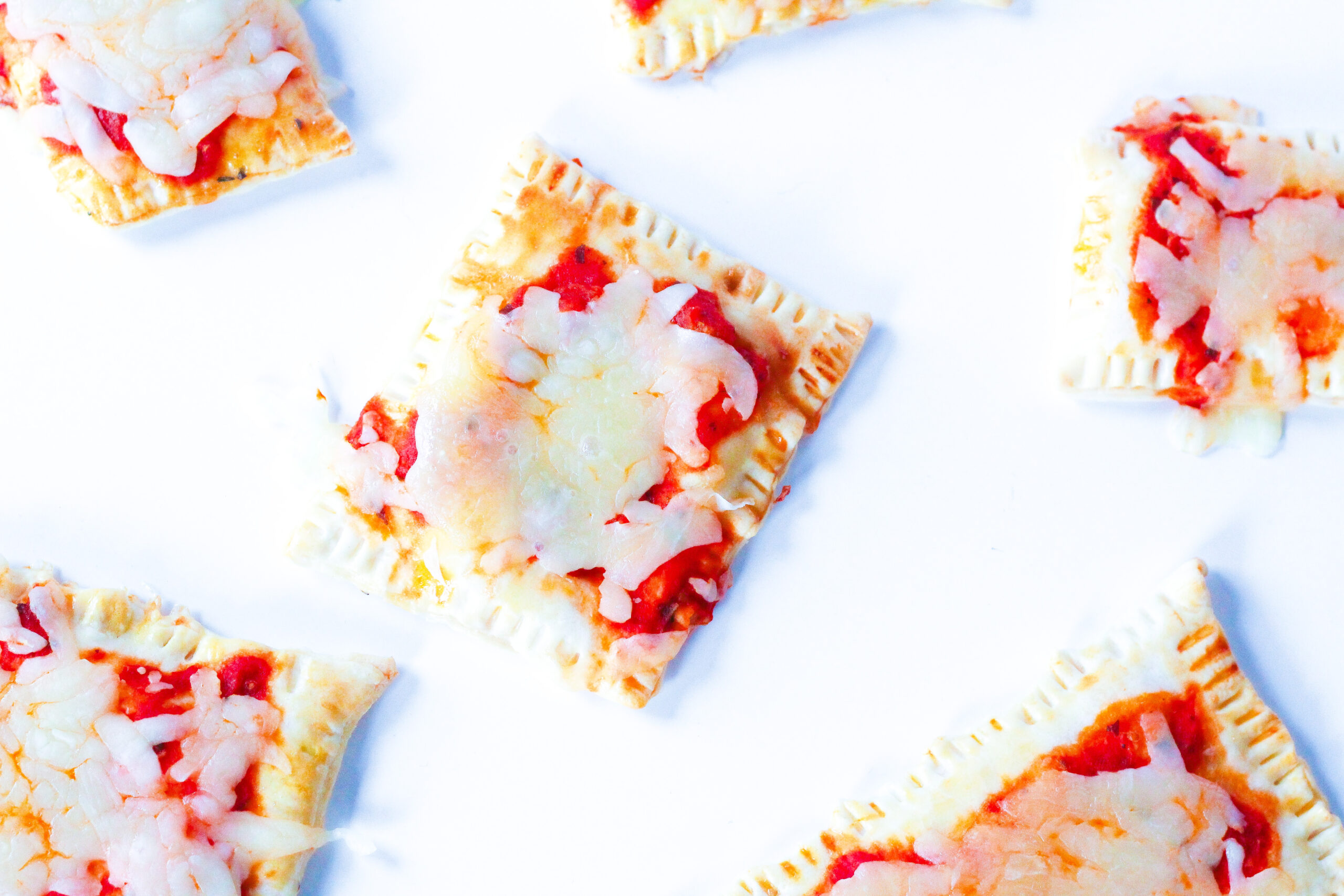 Top down view of pizza poptarts on a white surface. A square poptart is in the center, and all the poptarts surrounding it are partially out of the frame on all sides.