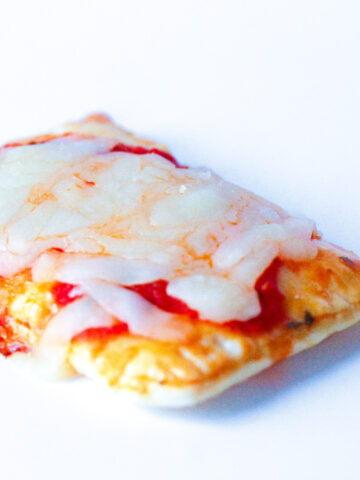Close up of a pizza poptart on a white surface. A golden flakey crust with fork imprints around the edges, topped with red pizza sauce and white mozzerella cheese