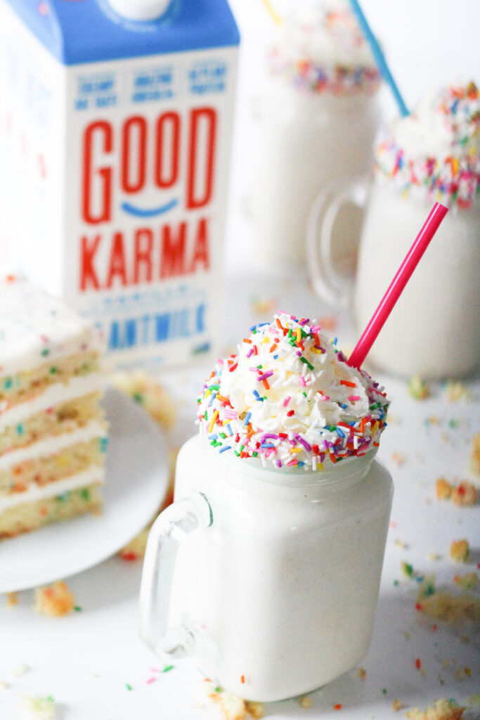 a birthday cake milkshake next to a giant slice of birthday cake with two more milkshakes and a milk carton in the background.