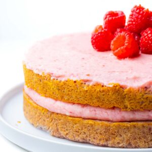 Two layers of raspberry cake going out of frame to the right with raspberry icing between the layers and on top and a small pile of raspberries on top.