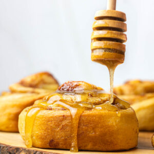 A wooden honey dipper drizzling honey onto an apple pie cinnamon roll.