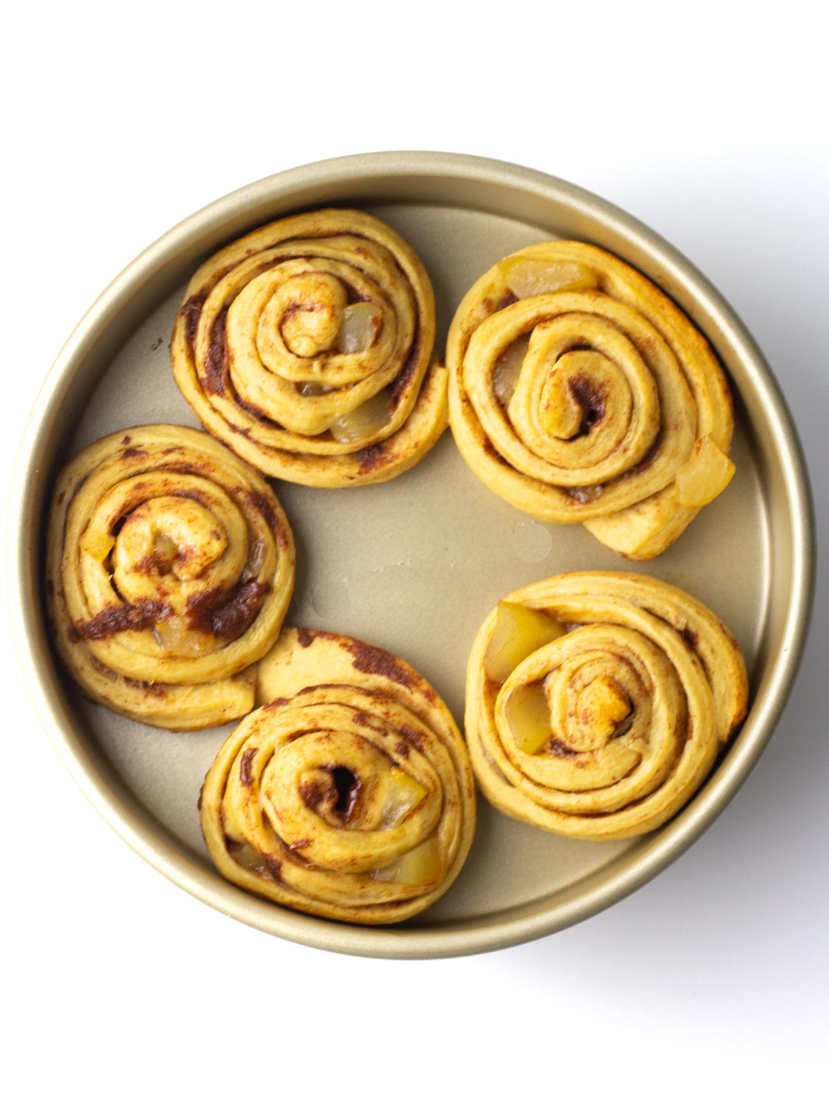 Five baked apple pie cinnamon rolls in a gold, round cake pan.