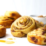 Close up of a cinnamon roll leaning on another cinnamon roll surrounded by more cinnamon rolls and a blurred bowl of honey glaze in the background.