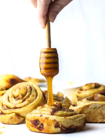 A hand holding a honey stick drizzling the honey brown sugar glaze onto a cinnamon roll surrounded by other cinnamon rolls.