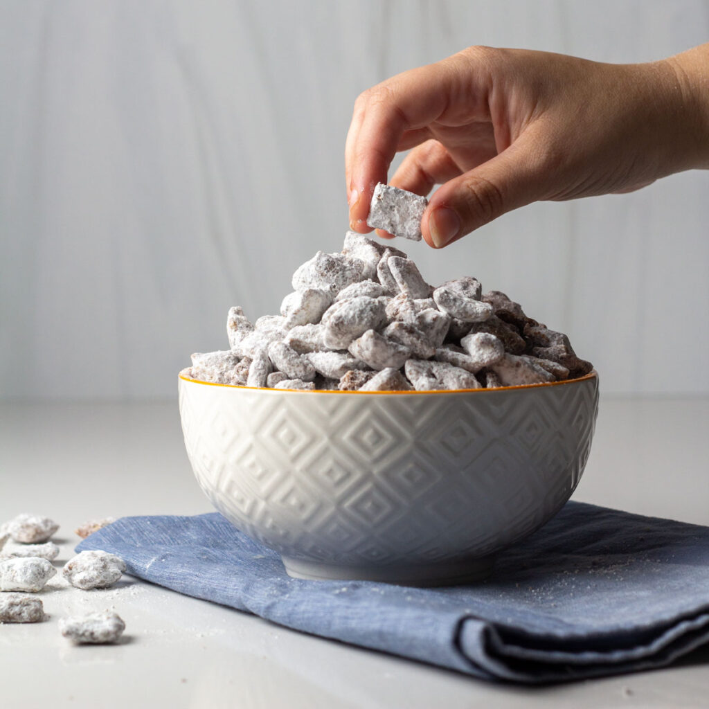 A hand reaching to a bowl of cinnamon toast crunch puppy chow sitting on a blue napkin.