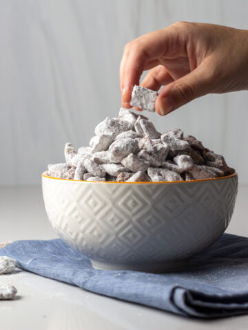A hand reaching to a bowl of cinnamon toast crunch puppy chow sitting on a blue napkin.