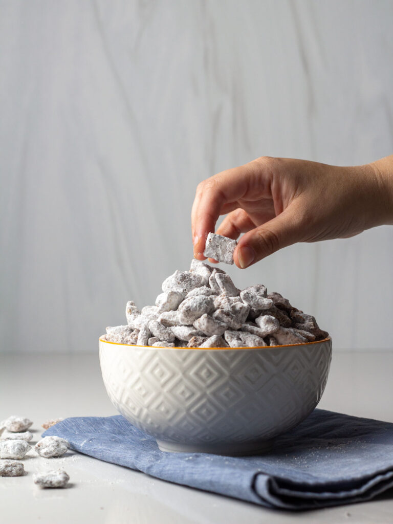 A hand reaching to a bowl of cinnamon toast crunch puppy chow sitting on a blue napkin.