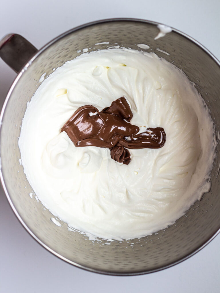 A mixing bowl of heavy whipping cream with soft peaks and Nutella on top.