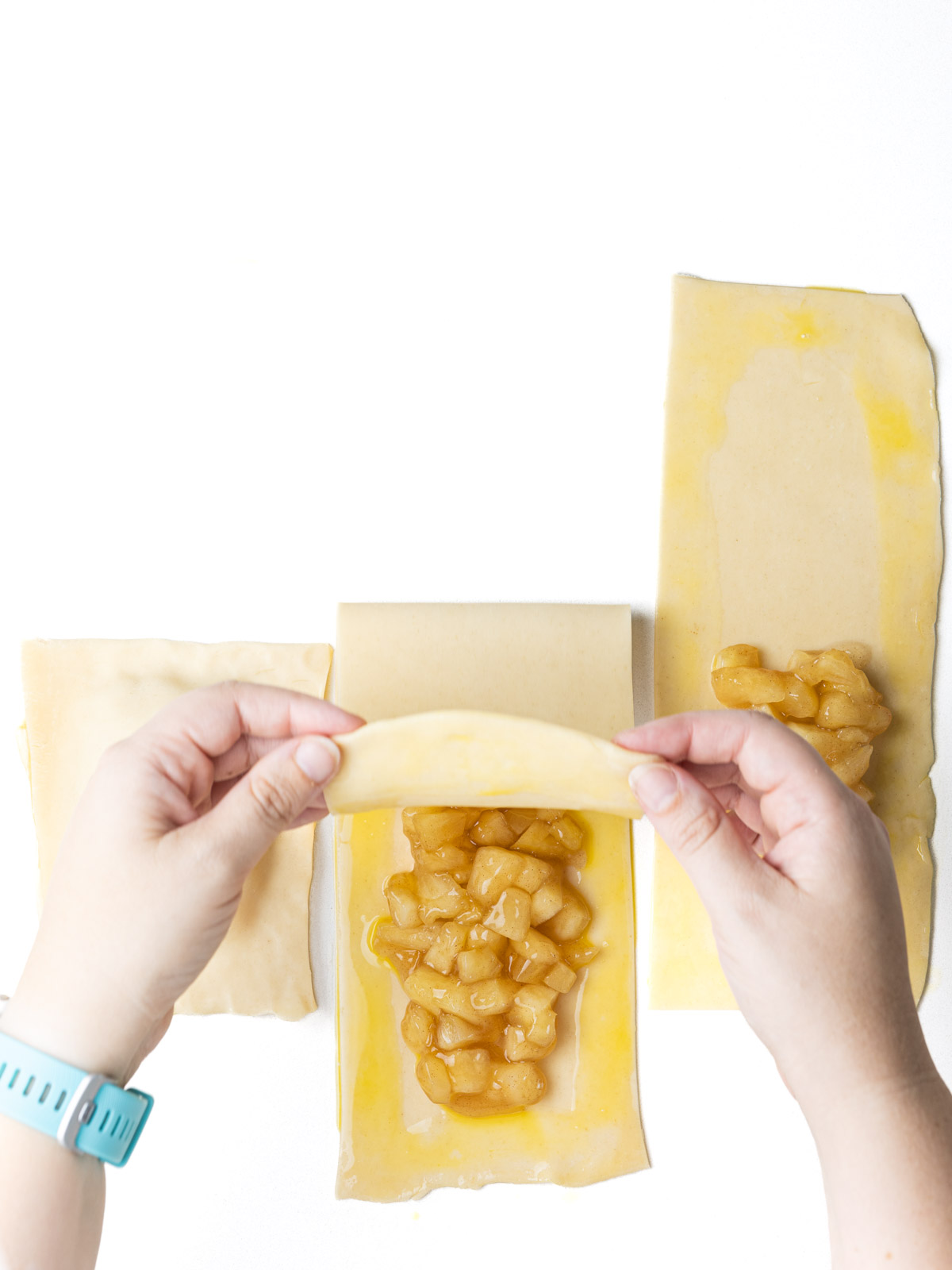 Hands folding half of the strip of pie crust over the half topped with apple pie filling.