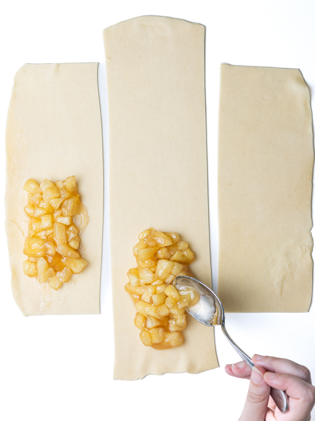 A hand spooning apple pie filling on to three rectangular strips of pie crust.