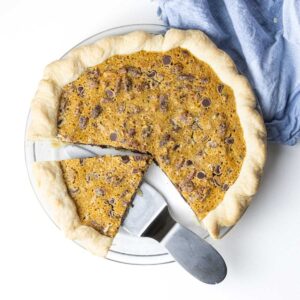 A top down view of a maple bourbon chocolate pecan pie with some pieces removed and one piece sliced with a pie server under it and a blue linen in the top right corner of the frame.