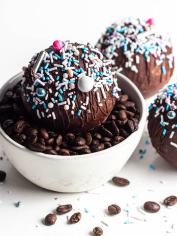 A mocha hot chocolate bomb covered in sprinkles sitting in a white bowl of coffee beans.
