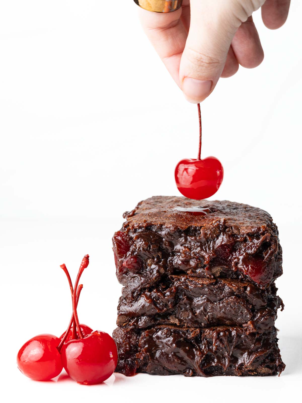 A hand holding a maraschino cherry over a stack of brownies with cherries next to the stack.