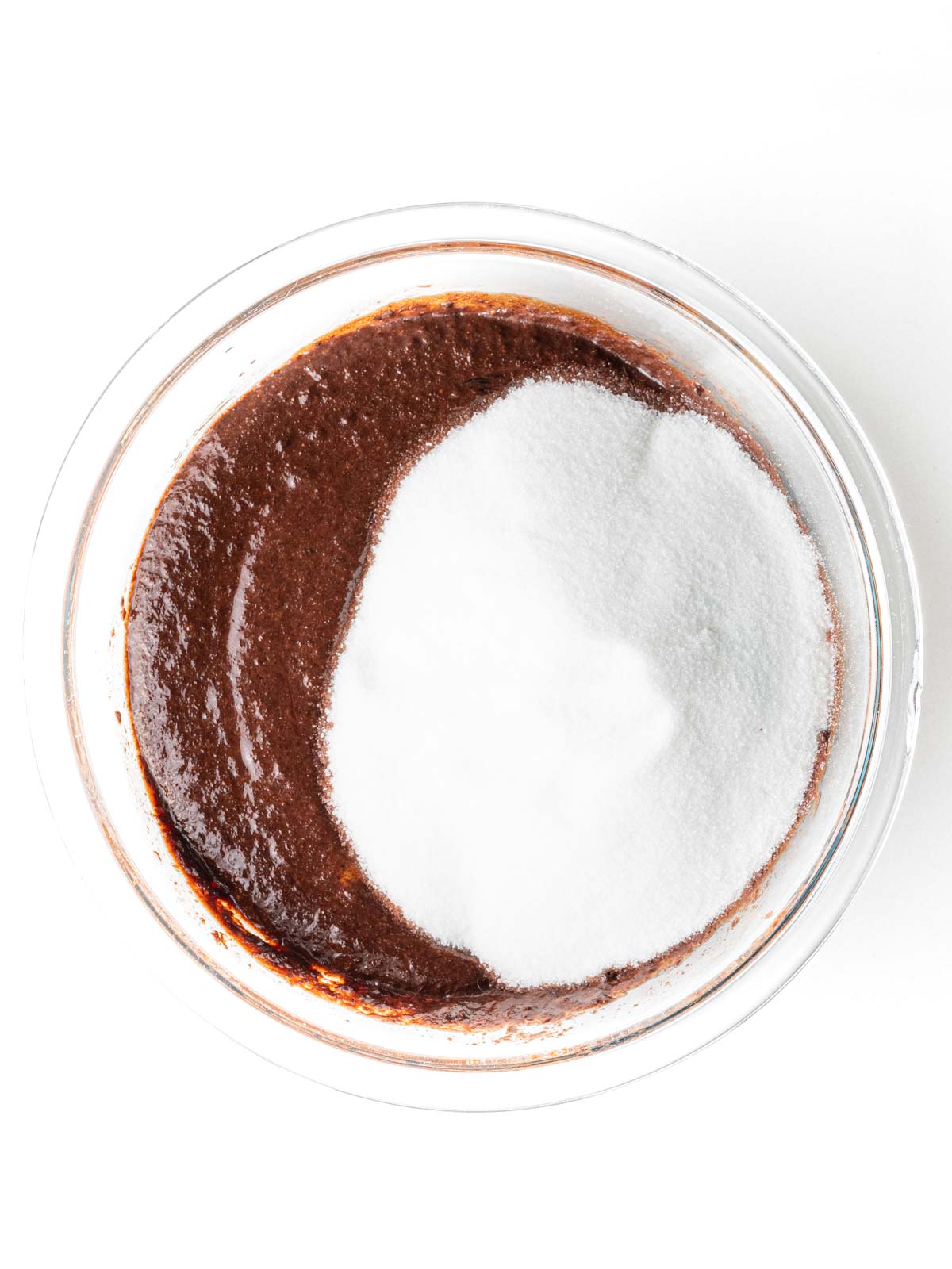 Sugar added to the cherry brownie batter in a clear glass mixing bowl.