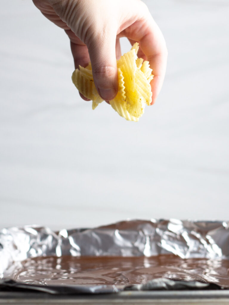 A hand holding broken potato chips over a chocolate lined sheet pan.