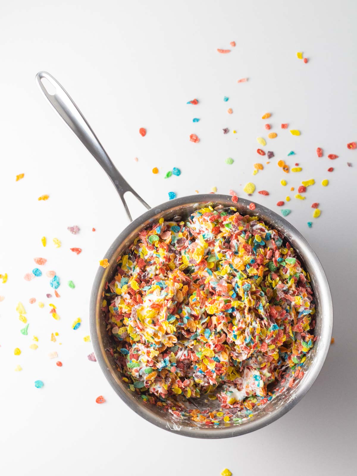 Fruity Pebbles stirred into melted marshmallow in a saucepan with fruity pebbles spilled around the outside of the pot.