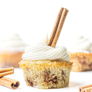 A cinnamon cupcake without a paper liner with a cinnamon stick sticking out of the frosting and a few cinnamon sticks on the surface next to the cupcake and some blurred cupcakes in the background.