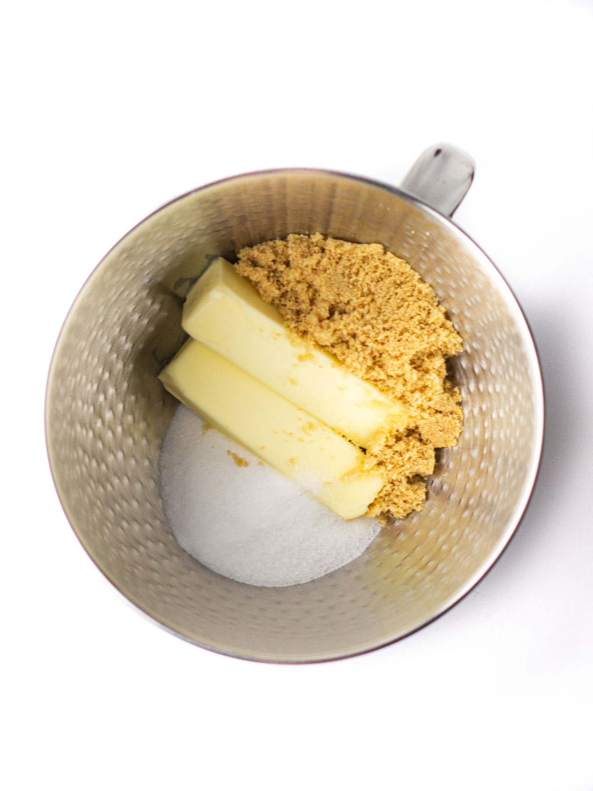 Two sticks of butter, sugar, and brown sugar in a silver textured mixing bowl.