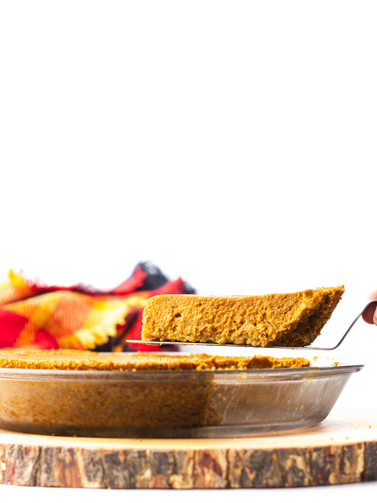 A slice of pumpkin pie being lifted out of the glass pie dish which is sitting on a round piece of wood.