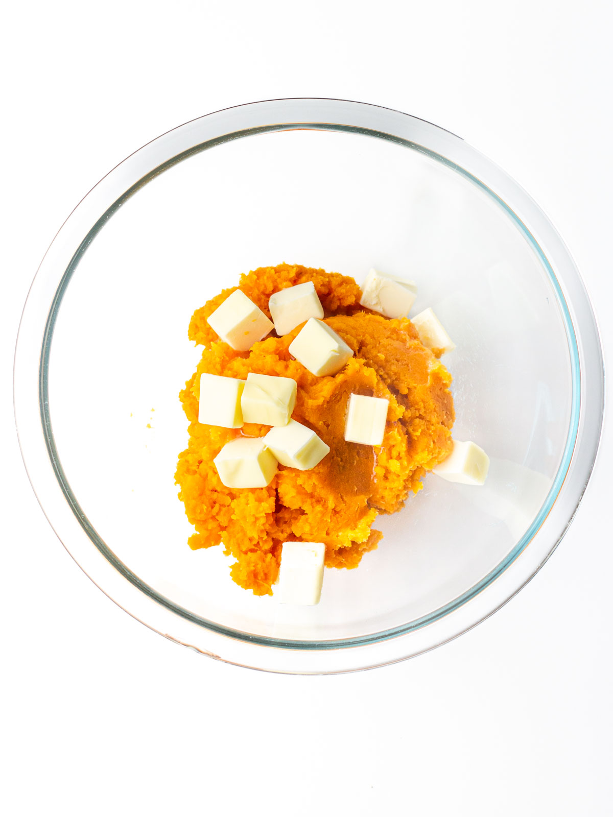 Sweet potato puree in a clear glass mixing bowl with cubes of butter on top.
