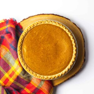 A whole fireball pumpkin pie on a round wooden plank with a red plaid cloth along the left side of the pie.
