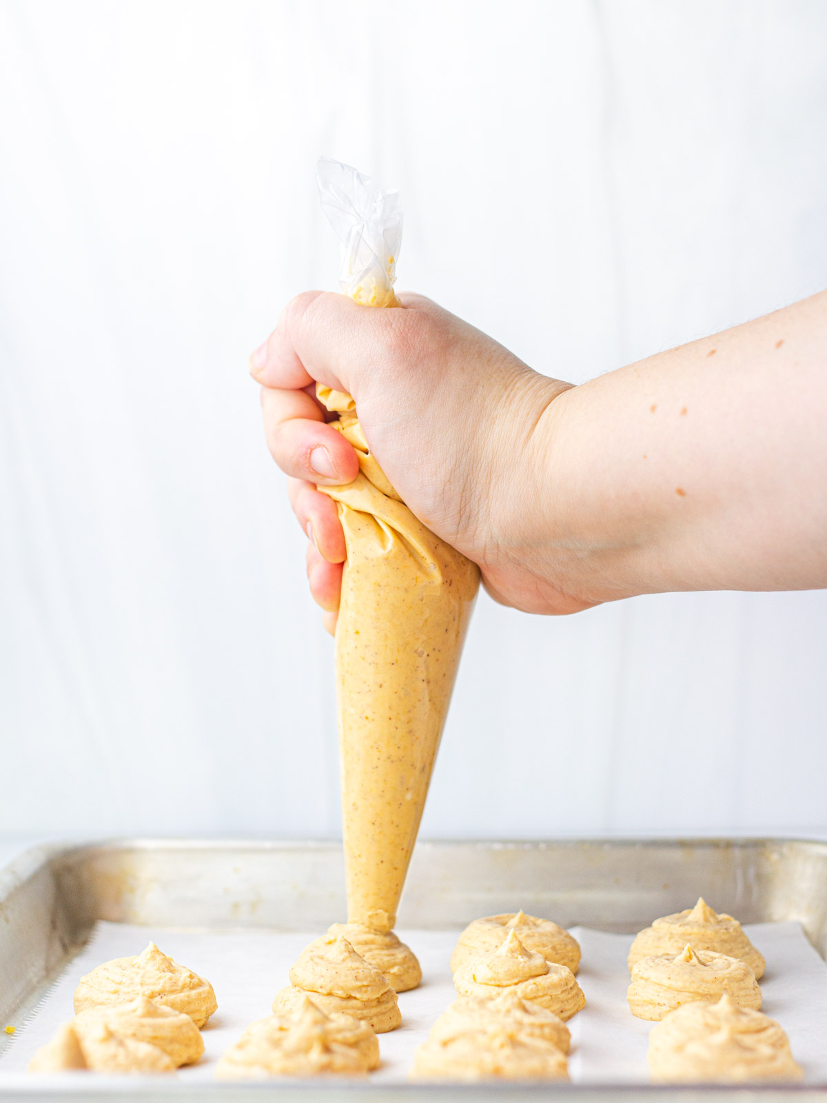 A hand holding a piping bag of pumpkin cheesecake filling piping dollops of filling onto a parchment lined baking sheet.