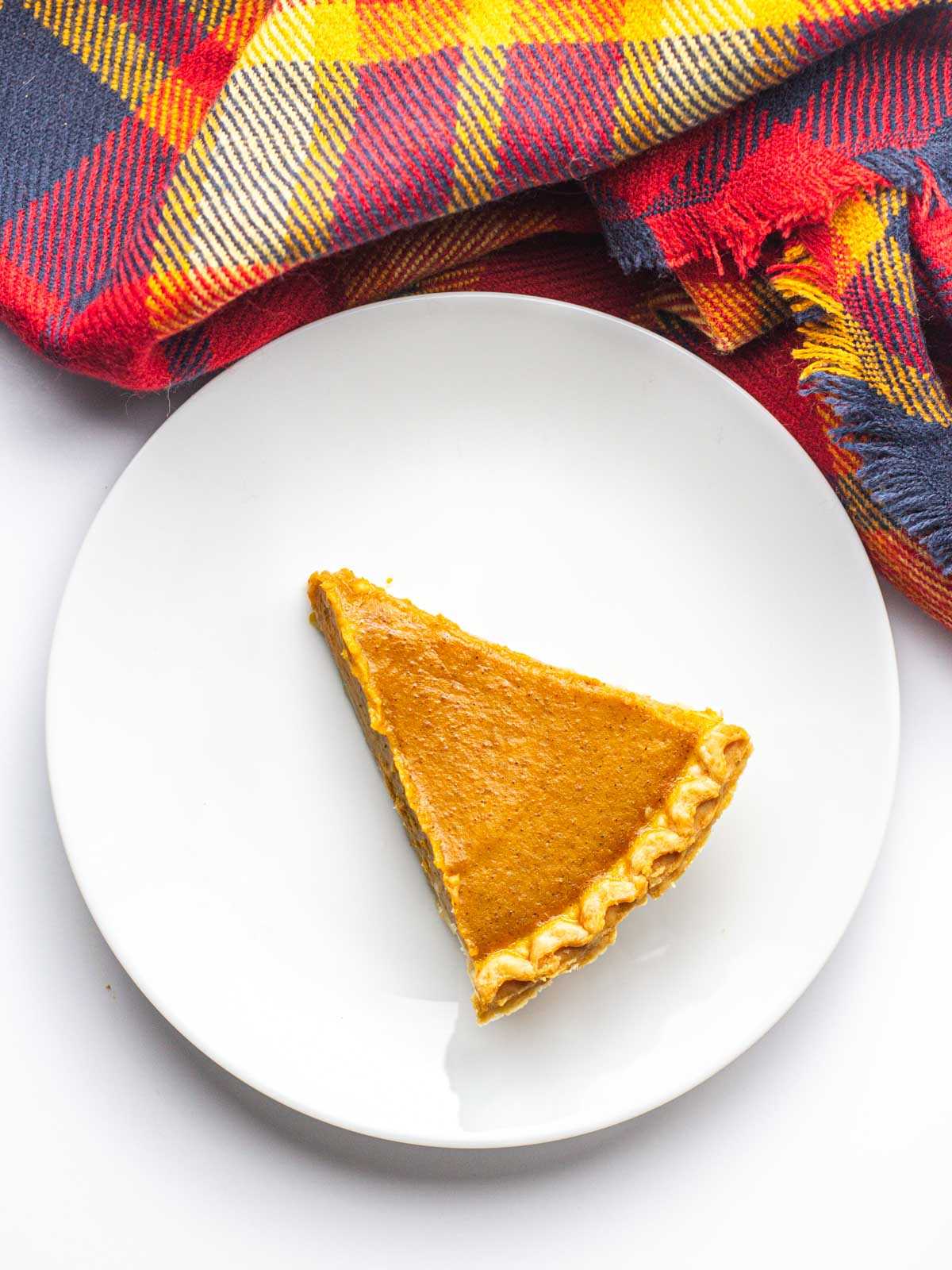 A slice of Fireball Pumpkin Pie on a round white plate with a red plaid cloth running along the top of the plate.