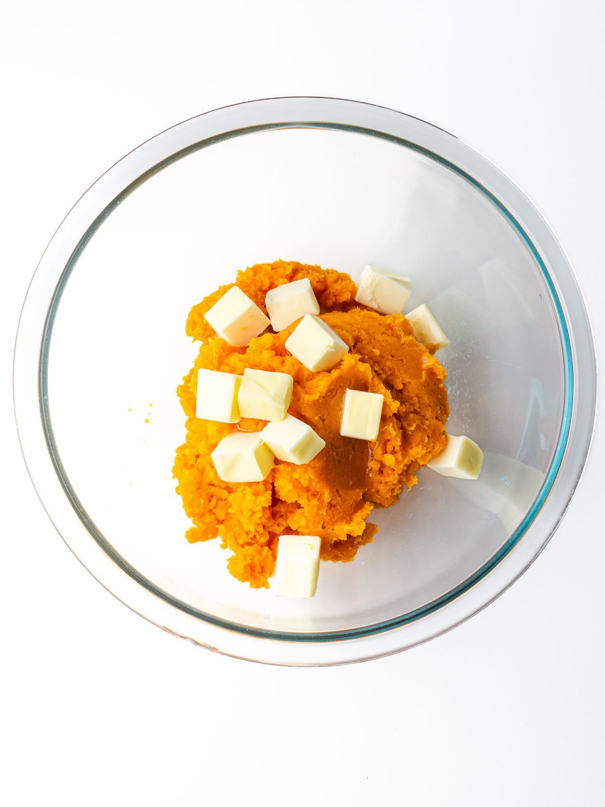 Sweet potato puree in a clear glass mixing bowl with cubes of butter on top.