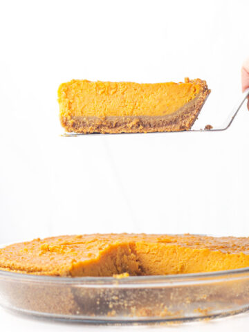 A hand holding a slice of sweet potato pie on a pie server over the rest of the pie.
