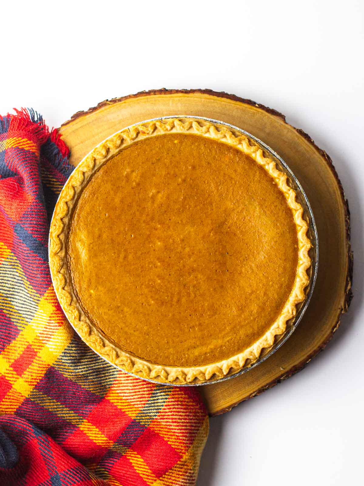 A whole fireball pumpkin pie on a round wooden plank with a red plaid cloth along the left side of the pie.