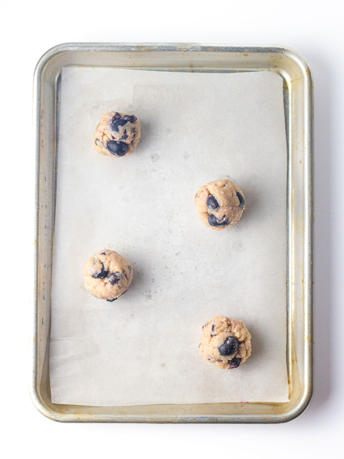Four blueberry cookie dough balls staggered on a parchment lined baking sheet.