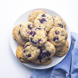 A batch of blueberry cookies on a round white plate placed on top of a blue napkin to the bottom right of the plate going out of frame.